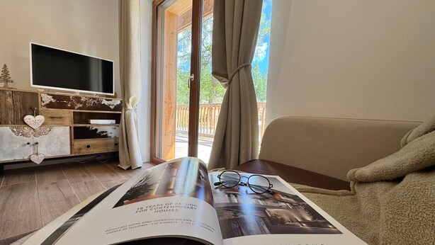 A room with a large window, a sofa, a magazine on a table, and a TV on a wooden cabinet.