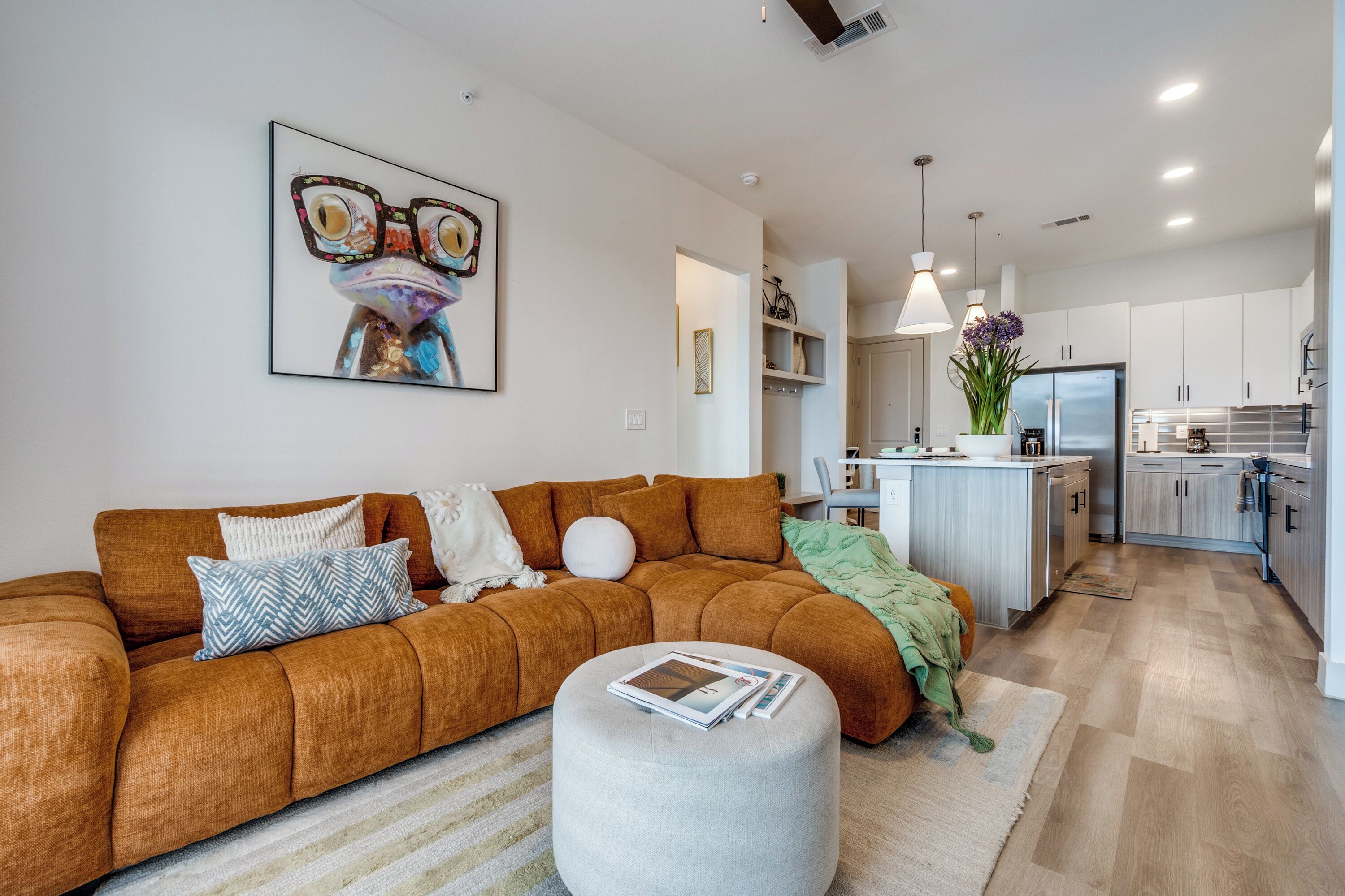 A modern living room with a brown sofa, a round ottoman, and a framed artwork on the wall.