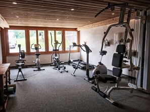 Ein Fitnessstudio mit verschiedenen Trainingsgeräten, großen Fenstern und einer Holzbalkendecke.