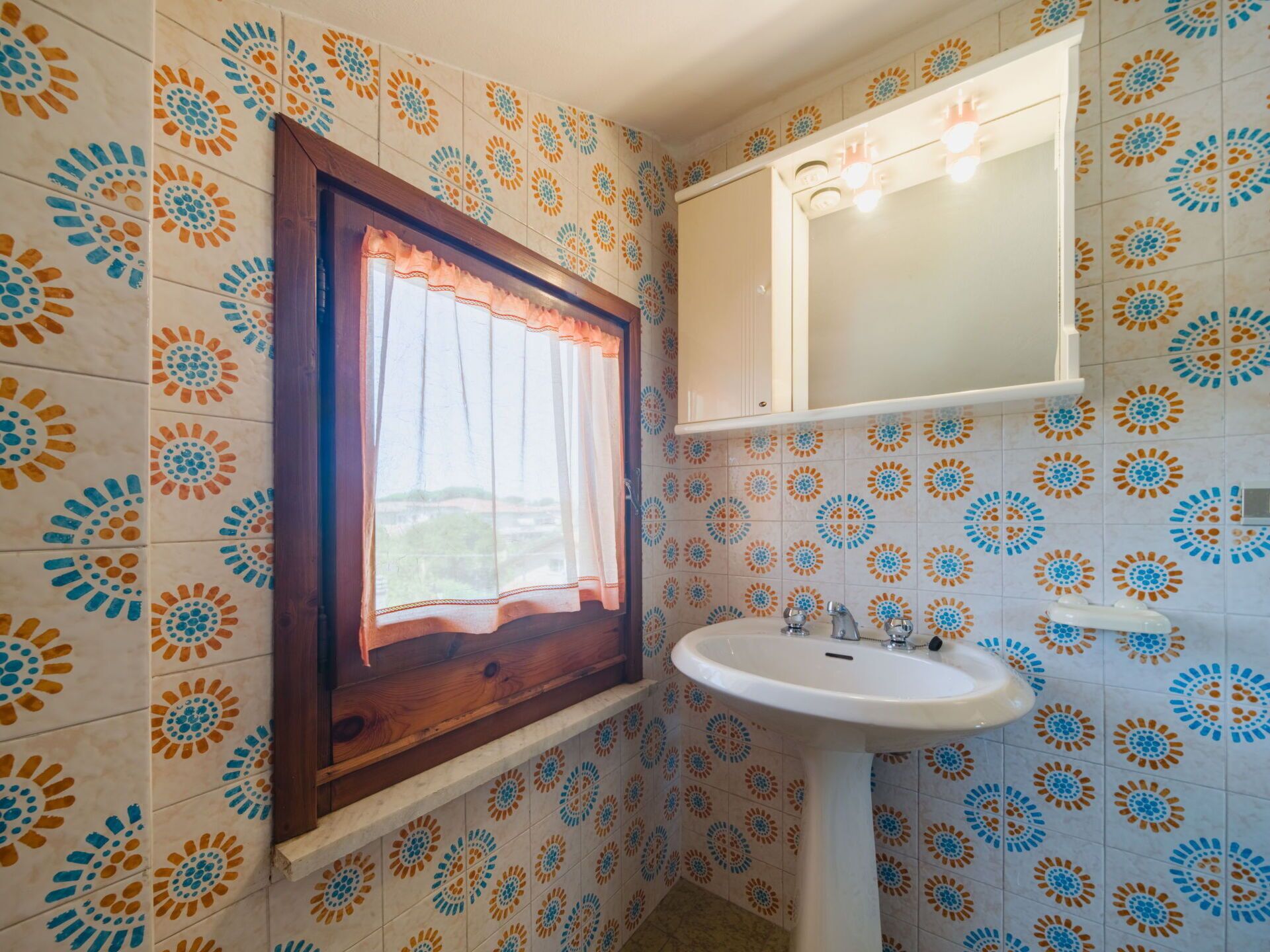 Une salle de bain avec des carreaux décorés, une fenêtre avec des rideaux, un lavabo et un miroir.