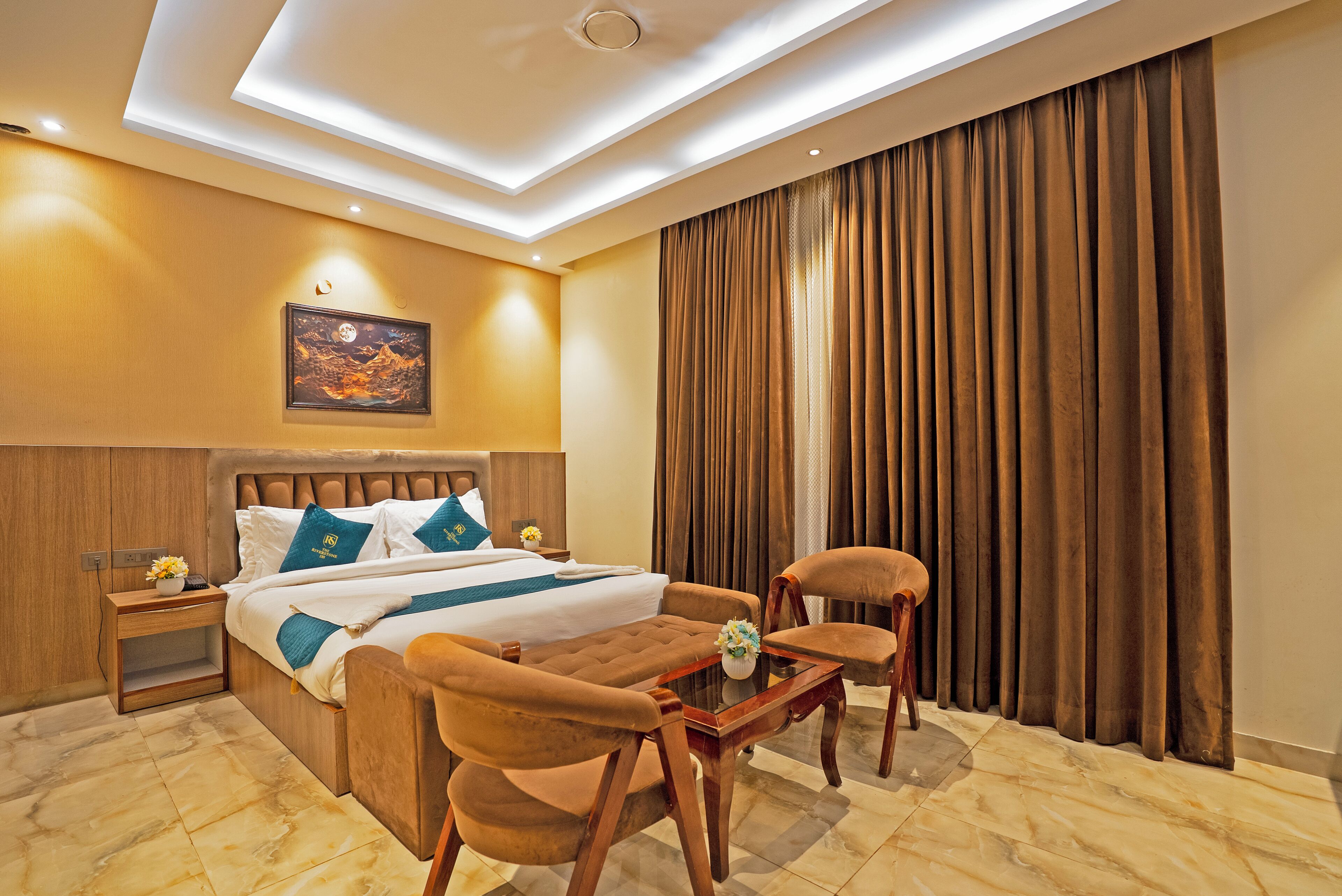 A hotel room with a large bed, a wooden nightstand, a desk, two chairs, and a large window with curtains.