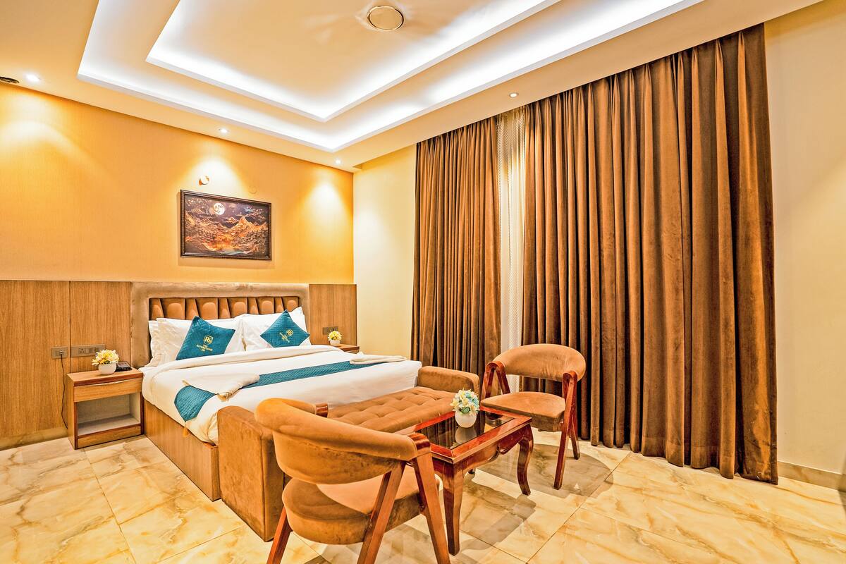 Habitación de hotel con cama, dos sillas, una mesita, mesita de noche, un cuadro y un ventanal grande con cortinas.
