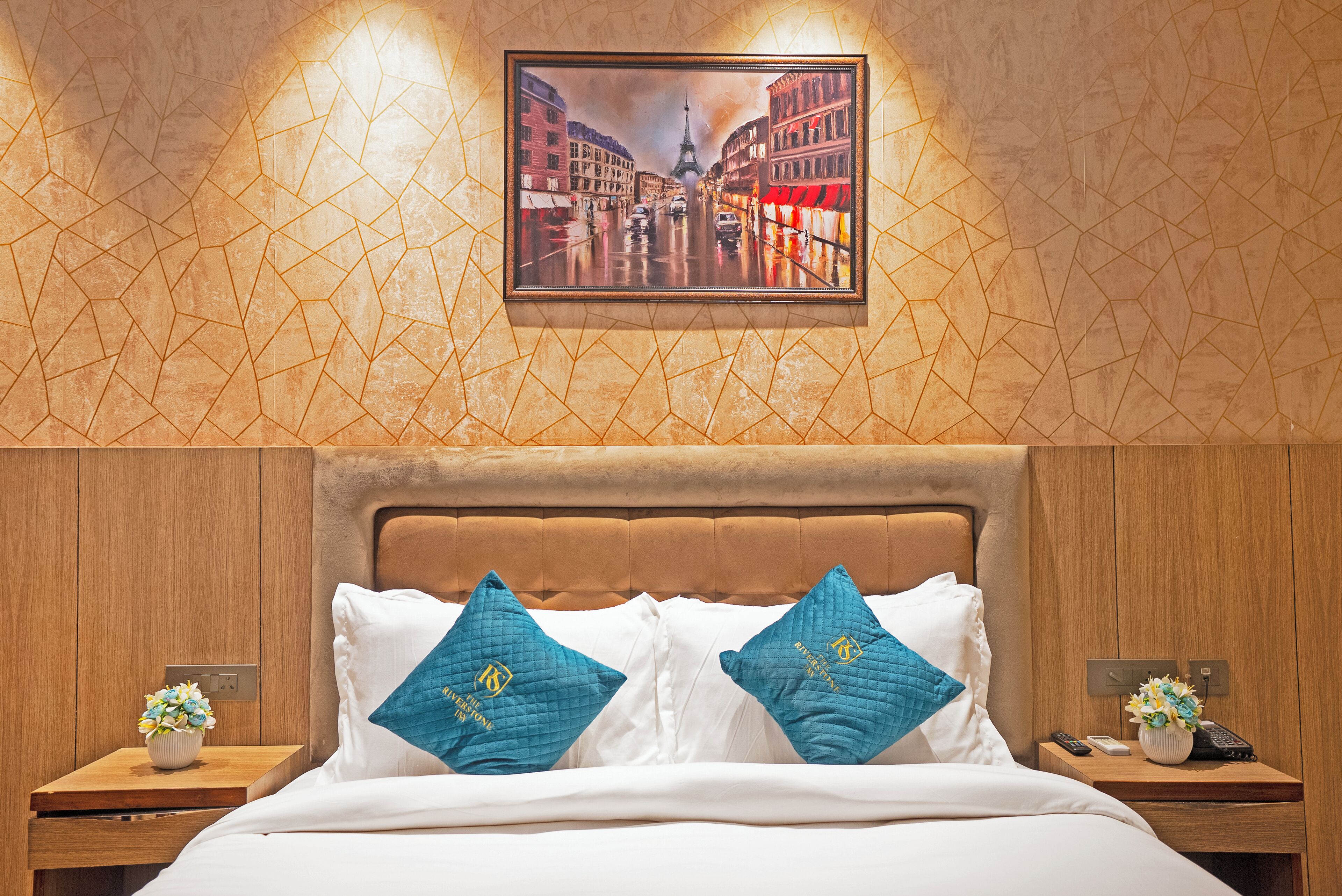Habitación de hotel con cabecera de madera, un cuadro enmarco que muestra una calle de la ciudad, dos mesitas de noche con jarrones de flores y dos almohadas color turquesa con un logotipo.