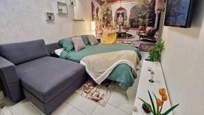 Ein modernes Wohnzimmer mit einer Couch, einem Bett und einem Esstisch. An den Wänden befinden sich Pflanzen und ein Gemälde.