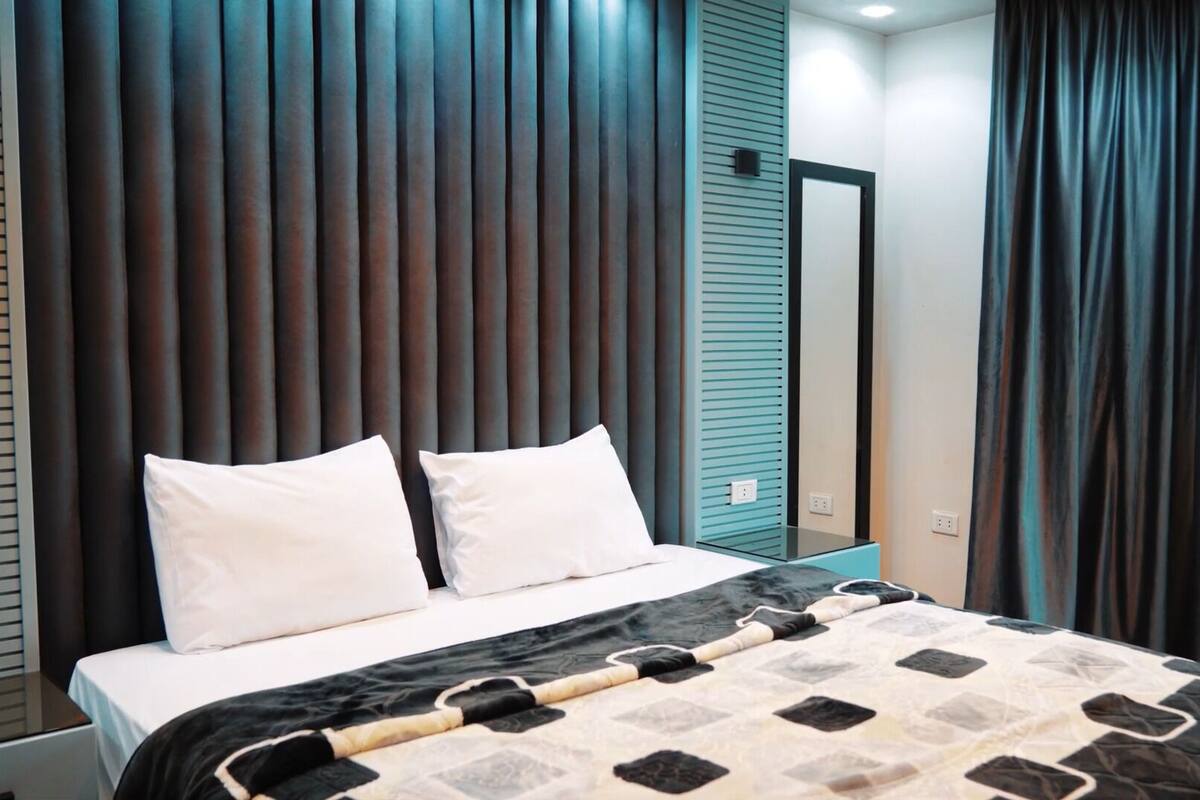 Un dormitorio moderno con una cama, dos almohadas, una colcha, una mesita de noche, un espejo y cortinas oscuras.