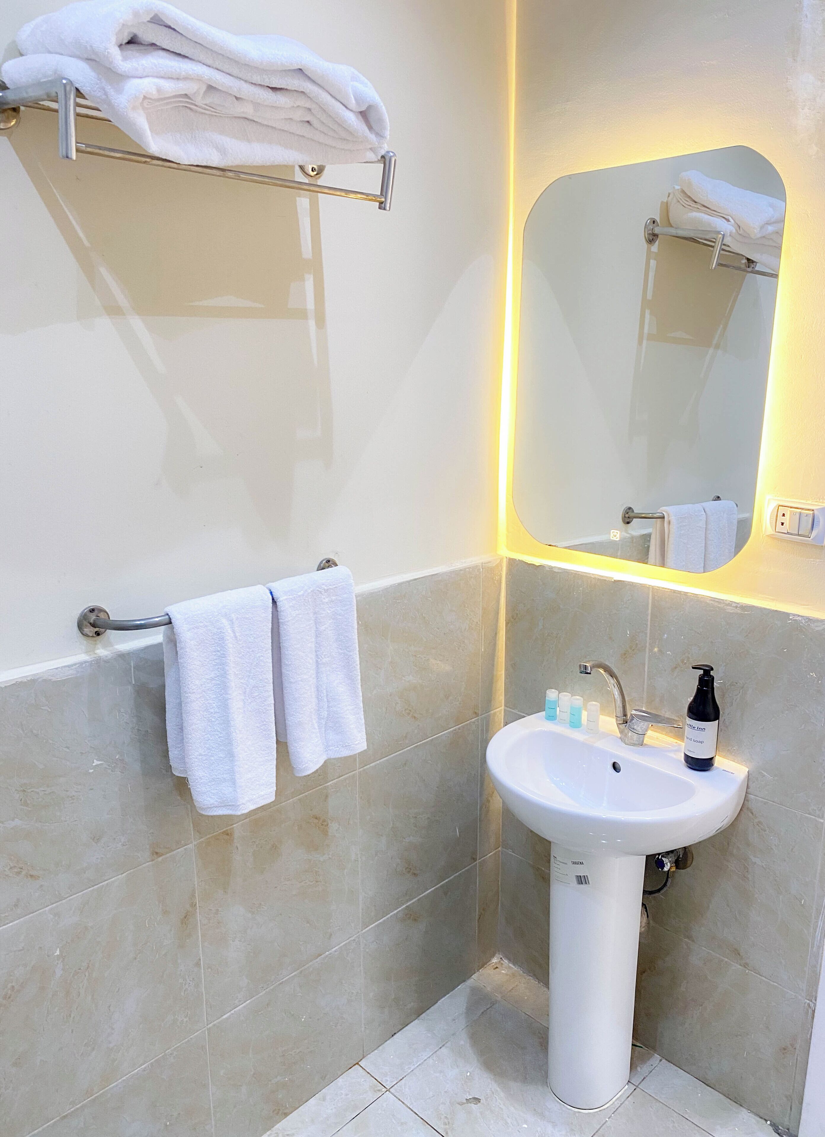 Un baño con un lavamanos blanco, dos toallas colgadas en un perchero, un espejo con luz y una botella de jabón sobre el lavamanos.