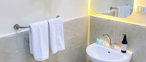 Un baño con un lavamanos blanco, dos toallas colgadas en un perchero, un espejo con luz y una botella de jabón sobre el lavamanos.