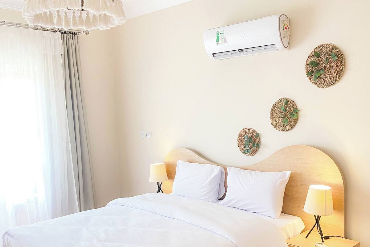 Un lit bien fait, avec du linge de lit blanc, dans une pièce au plancher en bois, dotée d’une lampe de chevet et d’un climatiseur fixé au mur.
