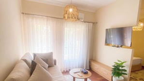 Ein Wohnzimmer mit einem beigefarbenen Sofa, einem kleinen runden Couchtisch, einem an der Wand montierten Fernseher und einer Topfpflanze.