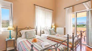 A bedroom with two beds, a window with a view, a radiator, and a balcony with a table and chair. - Villa Clio, with Mountain view (kalyves Armenoi)