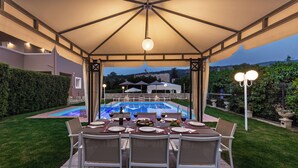 An outdoor dining area with a pool view, covered by a canopy, and surrounded by greenery. - Villa Clio, with Mountain view (kalyves Armenoi)