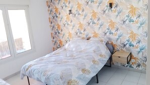 Un dormitori amb llit, una tauleta de nit, una finestra i una paret amb dibuix de fulles tropicals.