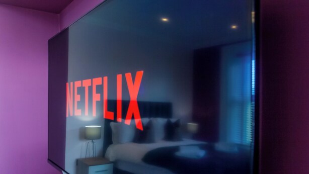 Quarto moderno com uma janela grande, uma cama, mesas de cabeceira e o logotipo da Netflix visível.