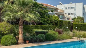 A swimming pool with tiled edges, surrounded by lush greenery and a palm tree. - Location Location Location - Garden Oasis in Limassol (Parekklisia)