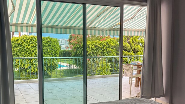 A hotel room with a bed, large sliding glass doors leading to a balcony with a view of greenery and a pool. - Location Location Location - Garden Oasis in Limassol (Parekklisia)