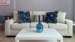 A living room with a light-colored sofa, two patterned throw pillows, a blue vase on a white coffee table, and a pink wall decoration. - Location Location Location - Garden Oasis in Limassol (Parekklisia)