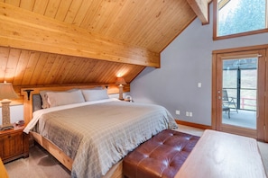 A bedroom with a wooden ceiling, a bed with a headboard, a bedside table with a lamp, and a window with a view of outdoor furniture.