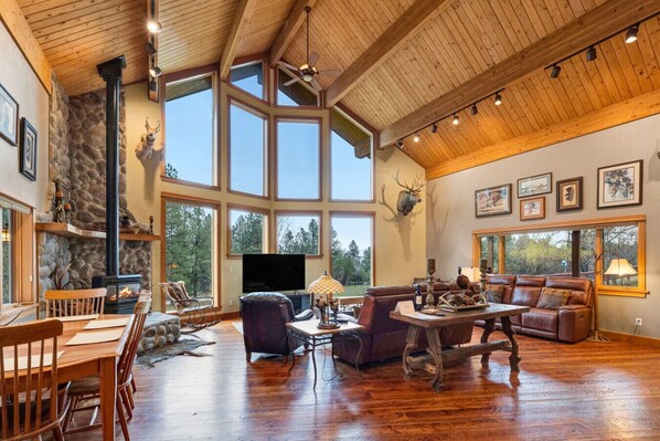 A spacious living room with a stone fireplace, large windows, and a high ceiling.