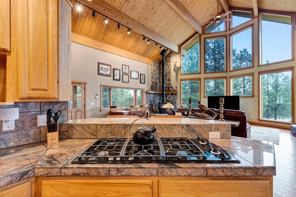 A spacious kitchen with granite countertops, a gas stove, wooden cabinets, and large windows offering a view of the outdoors.