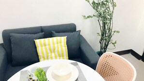 Un soggiorno moderno con un divano grigio, un cuscino a righe giallo e bianco, una piccola pianta in vaso, un cappello bianco su un piatto e un tavolo bianco con tovaglietta grigia.