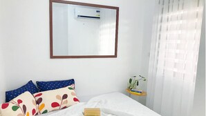 Un letto rifatto con biancheria bianca e cuscini colorati, un comodino con una pianta e una finestra con tende trasparenti.