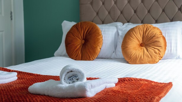 A neatly made bed with white linens, two orange pillows, and a rolled white towel on an orange blanket. - Modern Unit Cook  Work in Comfort (Motherwell)