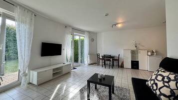 A modern living room with a flat-screen TV, a dining area with a table and chairs, a kitchen with a fridge and a coffee maker, and a large window with curtains.