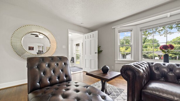 A living room with a tufted leather sofa, a round mirror with a gold frame, and a view of the outdoors through large windows. - Perfect Lindley Park location - Minutes from GAC, Coliseum, Restaurants and Pubs (Greensboro)