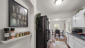 A modern kitchen with a black refrigerator, white cabinets, a herb garden wall art, and a dining area with a table and chairs. - Perfect Lindley Park location - Minutes from GAC, Coliseum, Restaurants and Pubs (Greensboro)