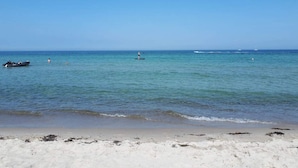 Una playa con aguas tranquilas, una orilla arenosa y algunos barcos a lo lejos.