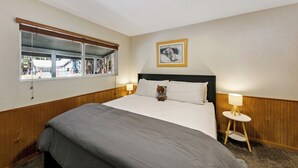 A hotel room with a large bed, a small bedside table with a lamp, a window with a view of the outside, and a framed picture on the wall.