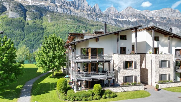 Un chalet de dos pisos con balcón, rodeado de exuberante vegetación y montañas.