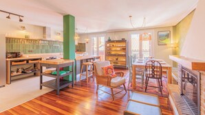 A spacious living area with a wooden floor, a dining table, chairs, a bookshelf, and a kitchen area with a green column.