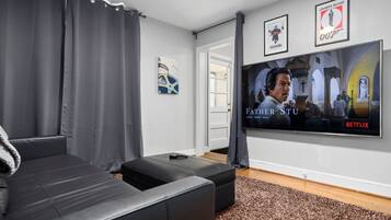 A living room with a large flat-screen TV displaying 'Father Stu' on Netflix, a black leather sofa, a grey curtain, and a framed picture of James Bond.