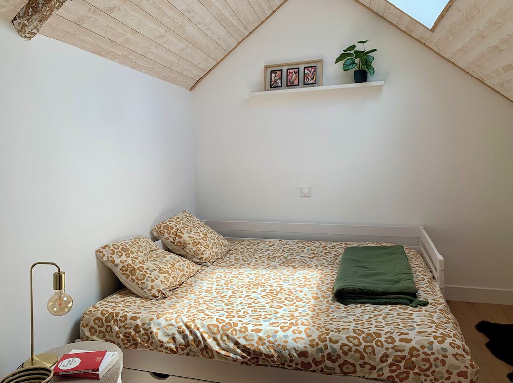 Un dormitorio con techo inclinado, una cama con ropa de cama de estampado de leopardo, una manta verde, una lámpara de noche y una pequeña estantería con fotografías y una planta.