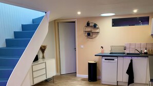 A modern kitchen with a built-in dishwasher, a dining area with a white table, and a blue staircase leading to another room. - Large duplex in the heart of the village of Carnac, near historic sites & beaches (Carnac)
