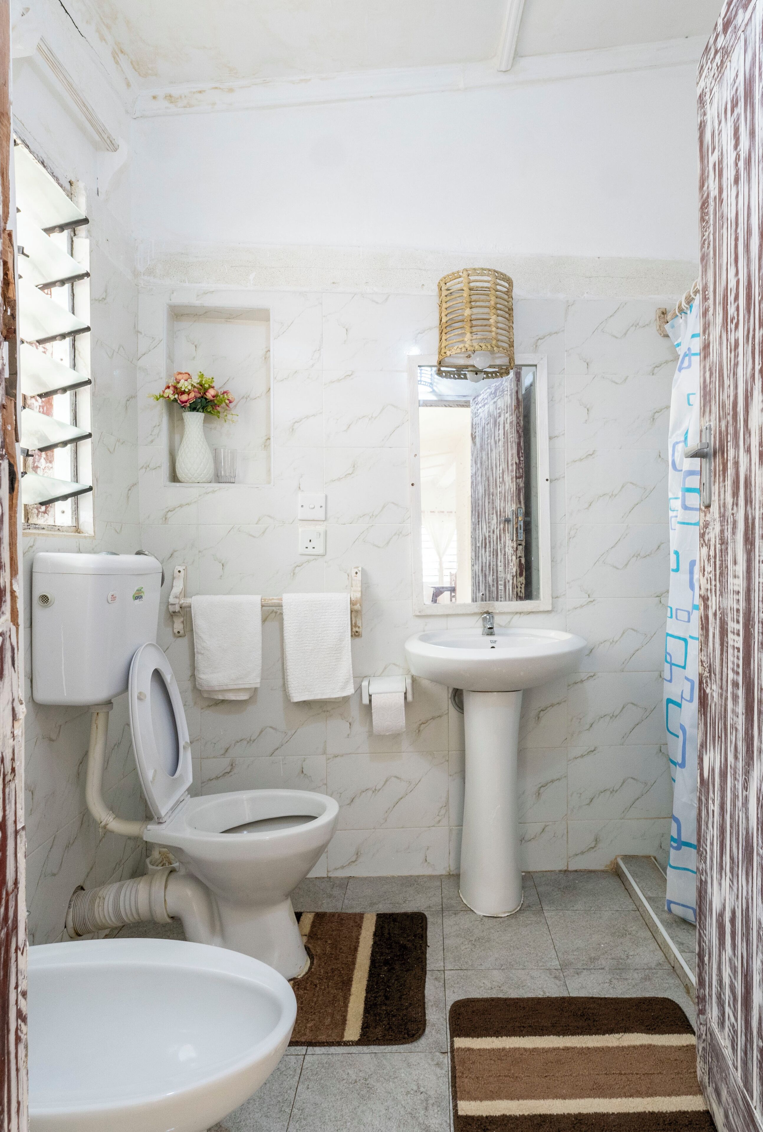 Une salle de bain comprenant des toilettes, un lavabo et un miroir. On y trouve un porte-serviettes avec des serviettes, une petite étagère avec un vase et un panier décoratif accroché au mur.