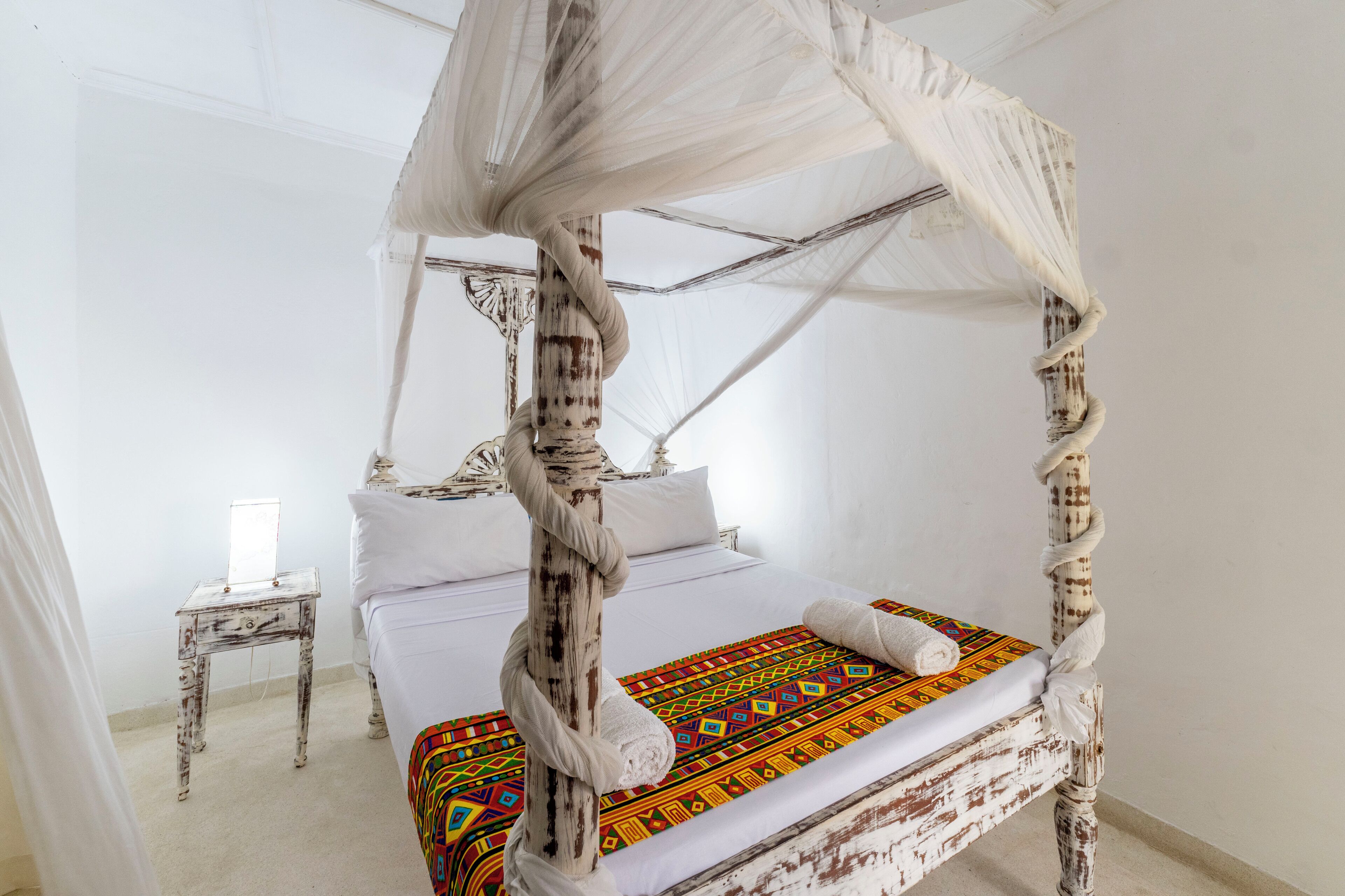 A four-poster bed with a colorful bedspread, a bedside table with a lamp, and a white curtain.
