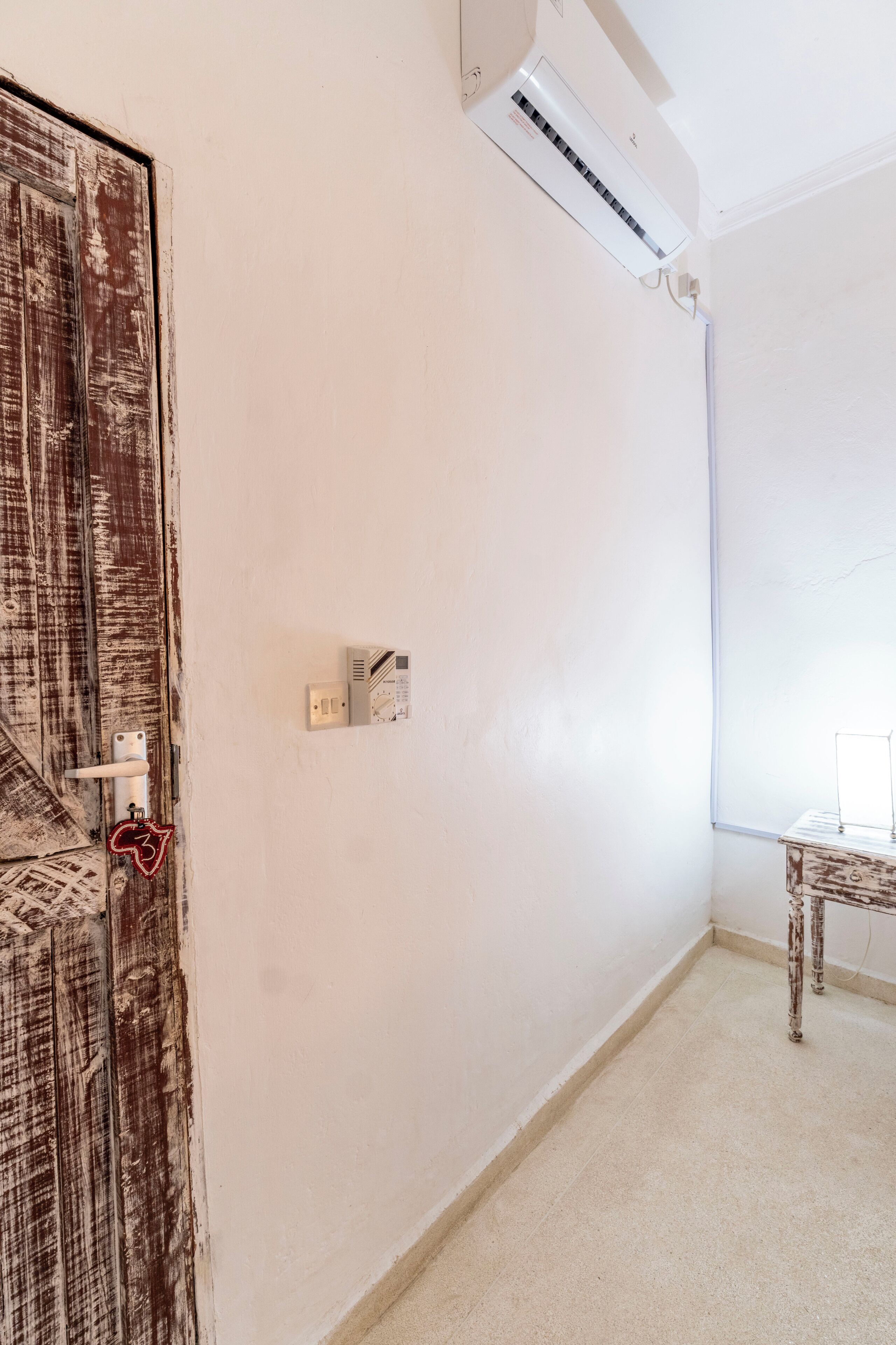 Une pièce avec un mur blanc, une porte en bois, un appareil de climatisation, un interrupteur et une petite table.