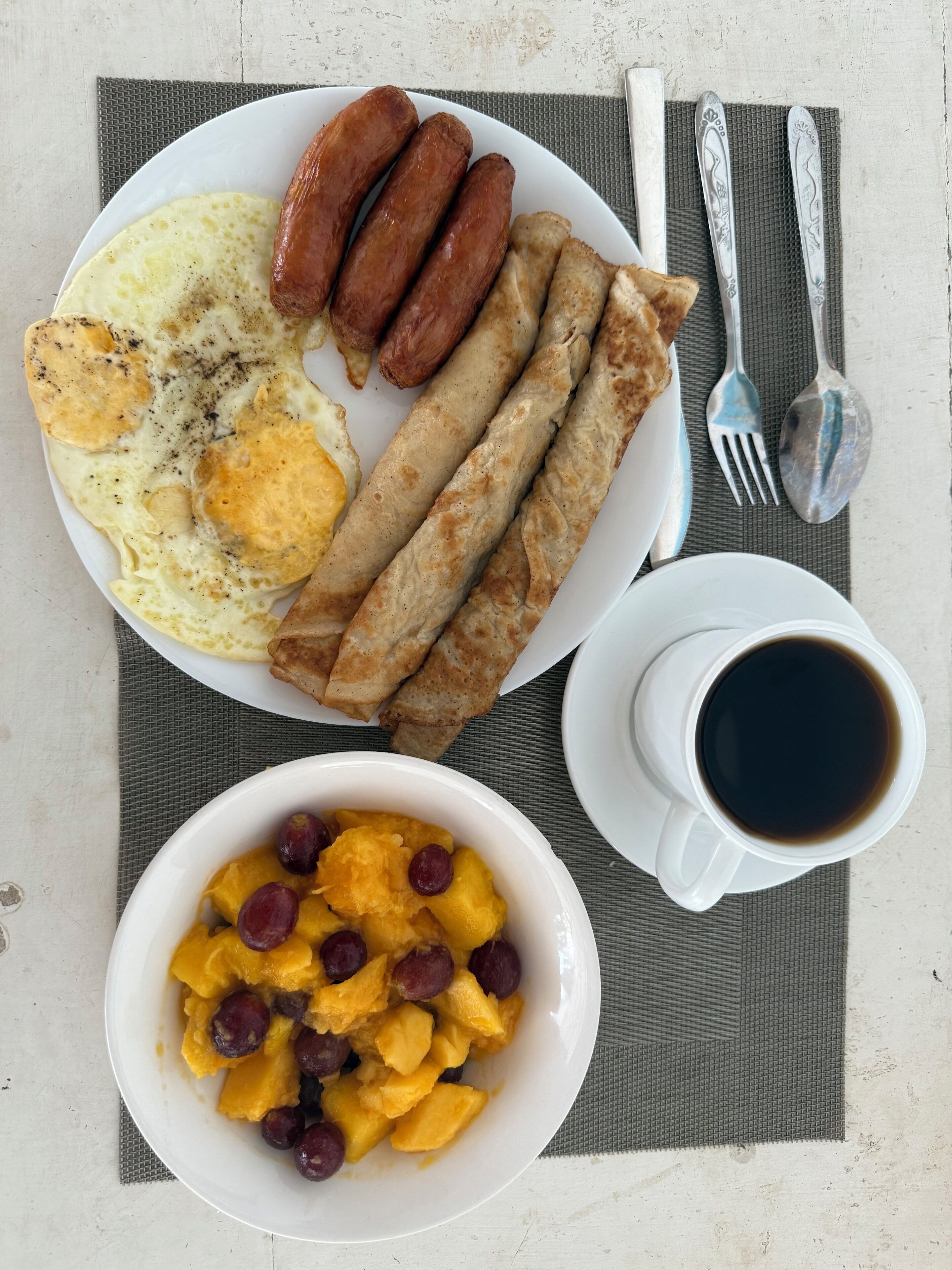 A breakfast plate with sausages, eggs, and pastries, a bowl of fruit, and a cup of coffee on a table.