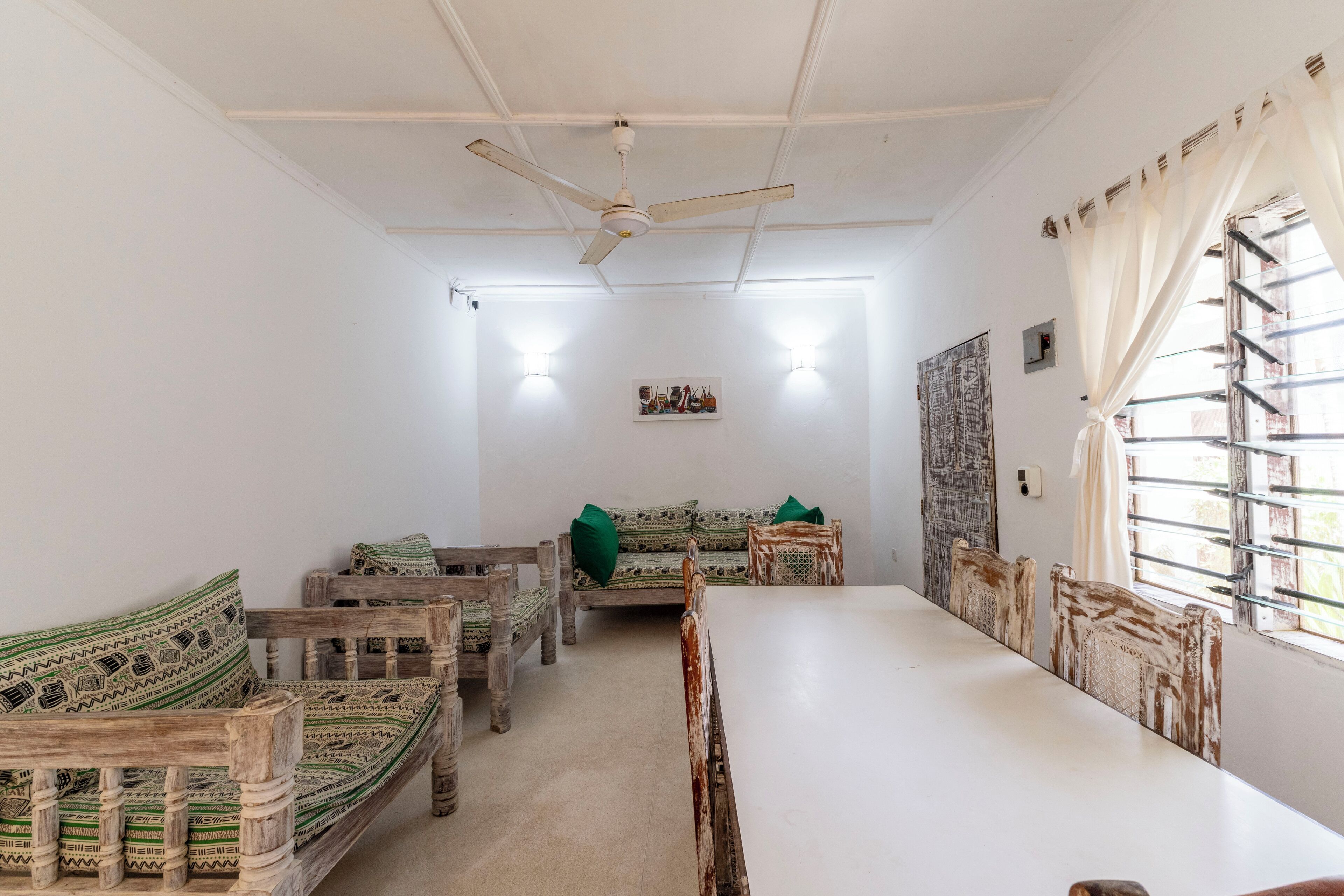 Una habitación con una mesa larga, sillas de madera, un sofá con cojines estampados, un ventilador de techo y una ventana con cortinas.