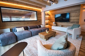 Een moderne woonkamer met een hoekbank, een witte fauteuil, een houten salontafel en een grote televisie bevestigd aan een houten wand.