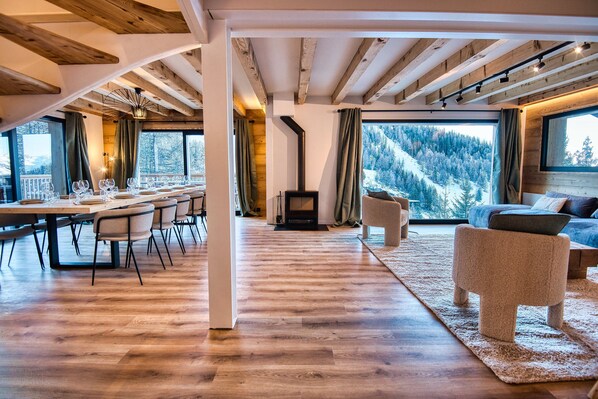 Un espacio habitable amplio con piso de madera, una mesa de comedor preparada para seis personas, una chimenea y un ventanal con vistas a una montaña nevada.