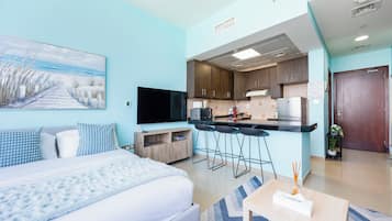 A modern hotel room with a bed, a TV, a kitchenette, and a painting on the wall.