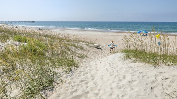 A sandy beach with a clear sky, a pier in the background, and a person walking towards the water. - Buddy's Sandcastle: room for family, friends, and pets. Fenced yard. (Atlantic Beach)