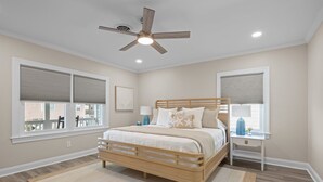 A bedroom with a bed, a nightstand, a ceiling fan, and large windows with blinds. - Buddy's Sandcastle: room for family, friends, and pets. Fenced yard. (Atlantic Beach)