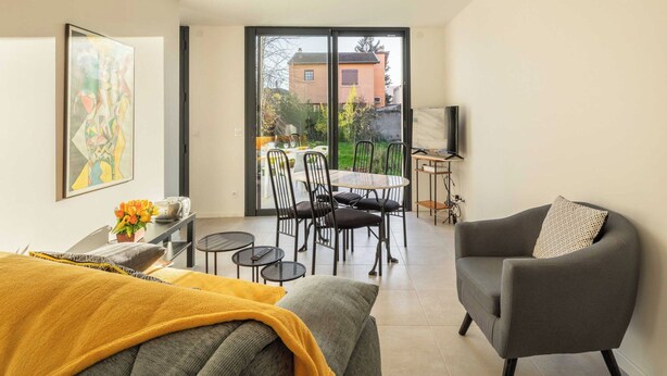 A modern living room with a sofa, a grey armchair, a dining table with chairs, and a large sliding glass door leading to an outdoor patio. - Dify Stoppa - Parilly (Venissieux)