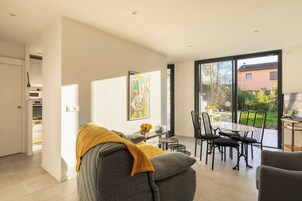 Una sala de estar moderna con un sofá, una mesa de centro y un comedor con sillas y una mesa. Hay un ventanal con vistas a un jardín y un edificio.