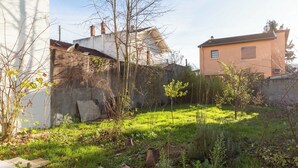 A garden with a variety of plants and a small tree. There is a white wall and a building with a brown roof in the background. - Dify Stoppa - Parilly (Venissieux)