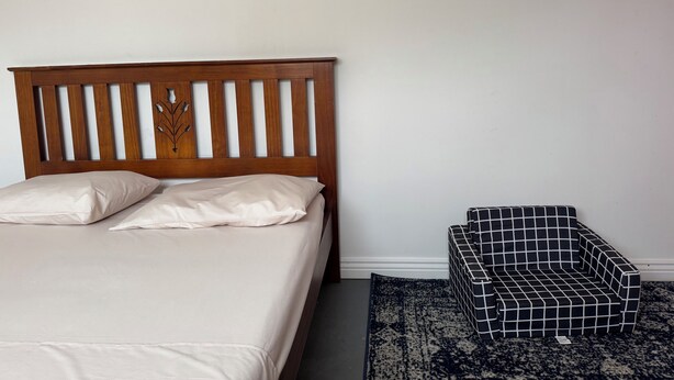 A bed with a wooden headboard, a black and white checkered ottoman, and a dark patterned rug. - A cosy sleep out in backyard (Kennington)